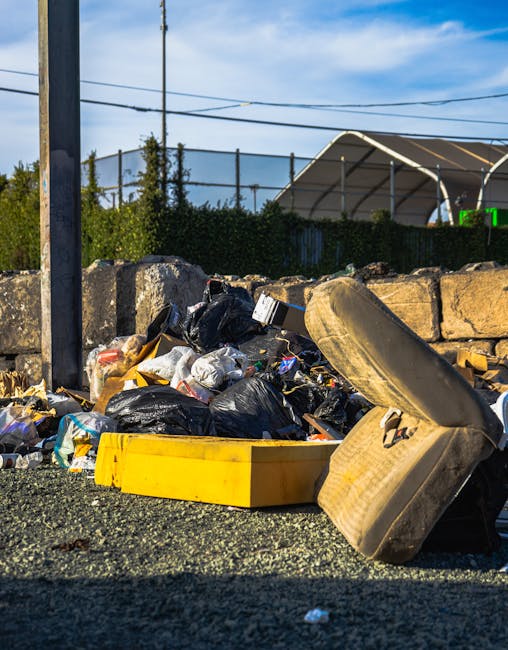 A pile of various discarded cardboard boxes, some flattened and others partially opened, stacked haphazardly against a rough concrete wall in an outdoor setting. Surrounding the boxes are crumpled pap